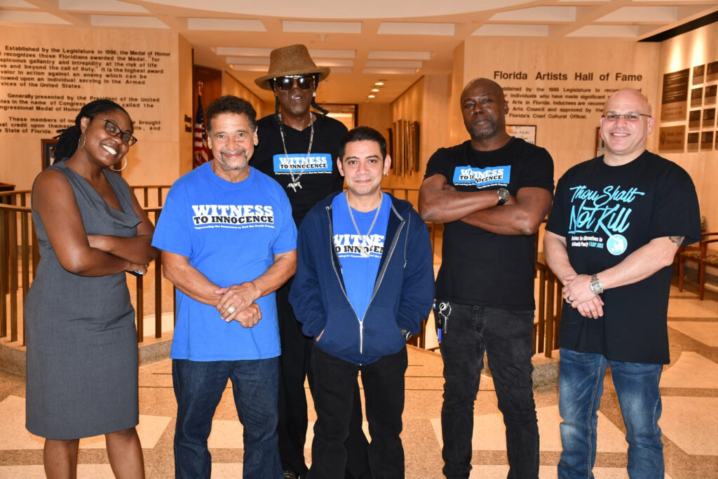 A group of five men and one woman stand in a state capitol building. The men wear black and blue t-shirts that say "Witness to Innocence". Clemente Aguirre is in the center. 